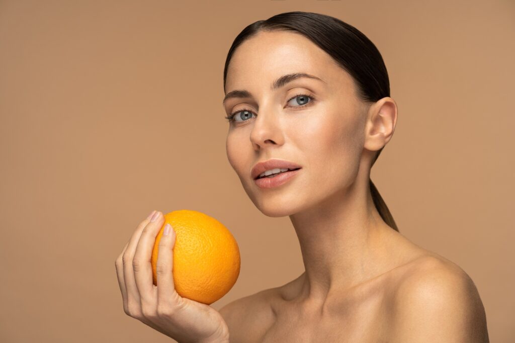 Woman with perfect face skin, natural makeup holding orange fruit over beige studio. Vitamin C