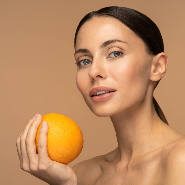 Woman with perfect face skin, natural makeup holding orange fruit over beige studio. Vitamin C